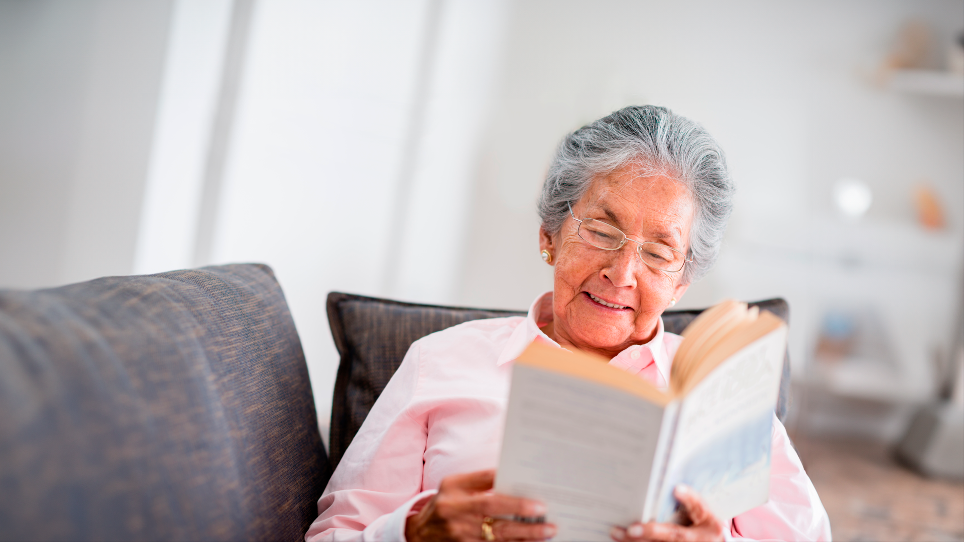 Mujer adulta mayor sonriente lee libro sentada en el sofá de la sala de sus casa. Día de la persona adulta mayor 2024.