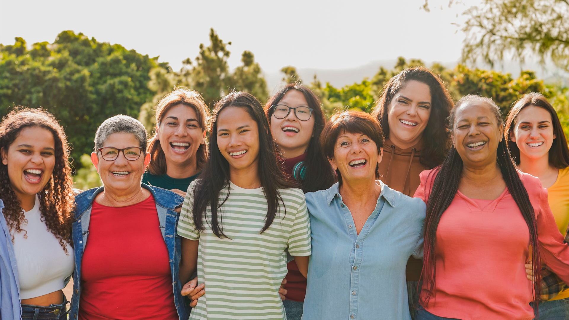 Grupo de mujeres de diversas edades y etnias posan en fila, sonrientes frente a la cámara en día soleado. Día Internacional de la Mujer 2025.