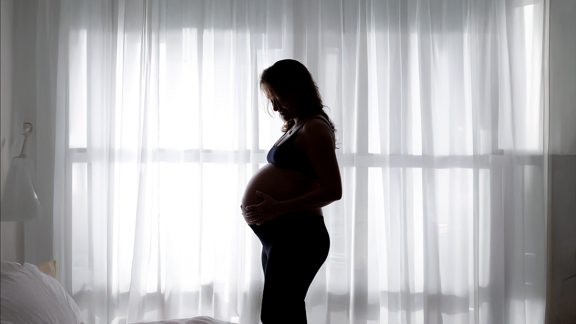Imagen ilustrativa. Mujer embarazada, de pie a contraluz frente a la ventana de su habitación.
