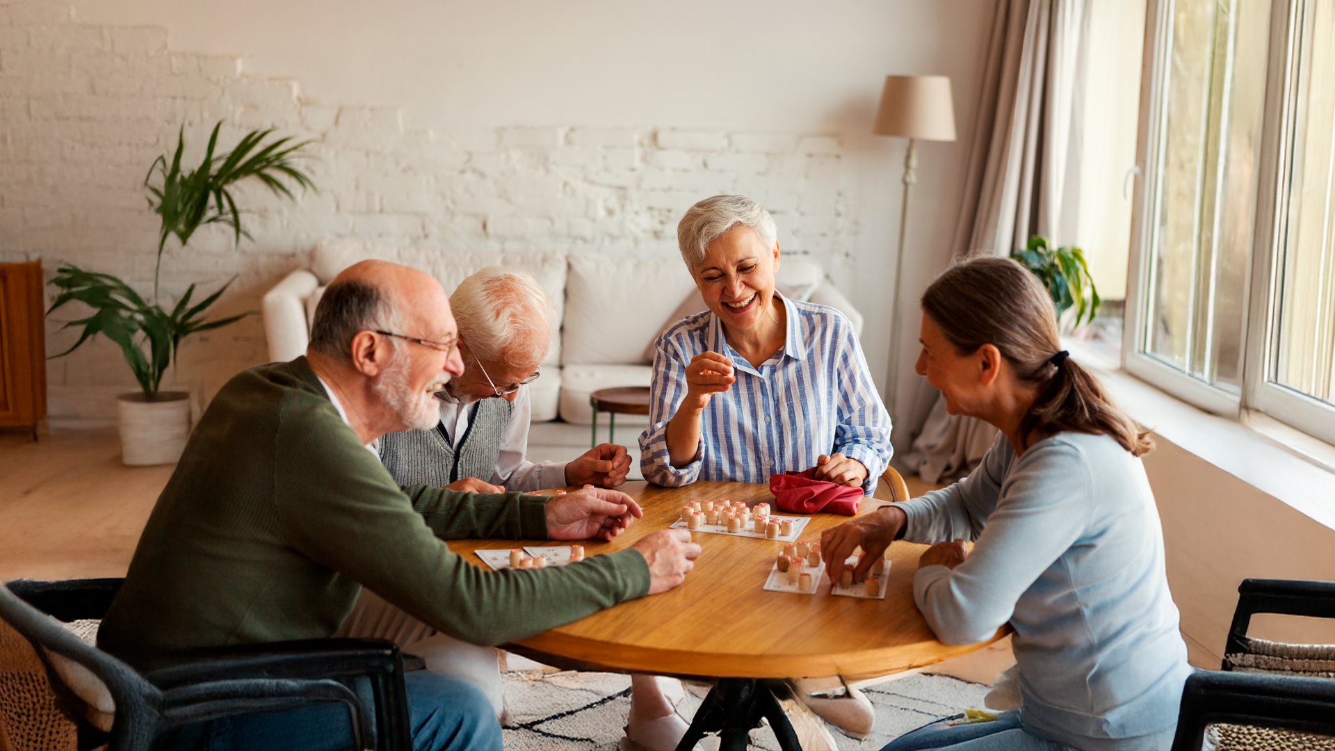 Imagen ilustrativa. Cuatro personas adultas mayores sonríen sentadas al rededor de una mesa mientras juegan. Especial del Día de la Persona Adulta Mayor 2025.