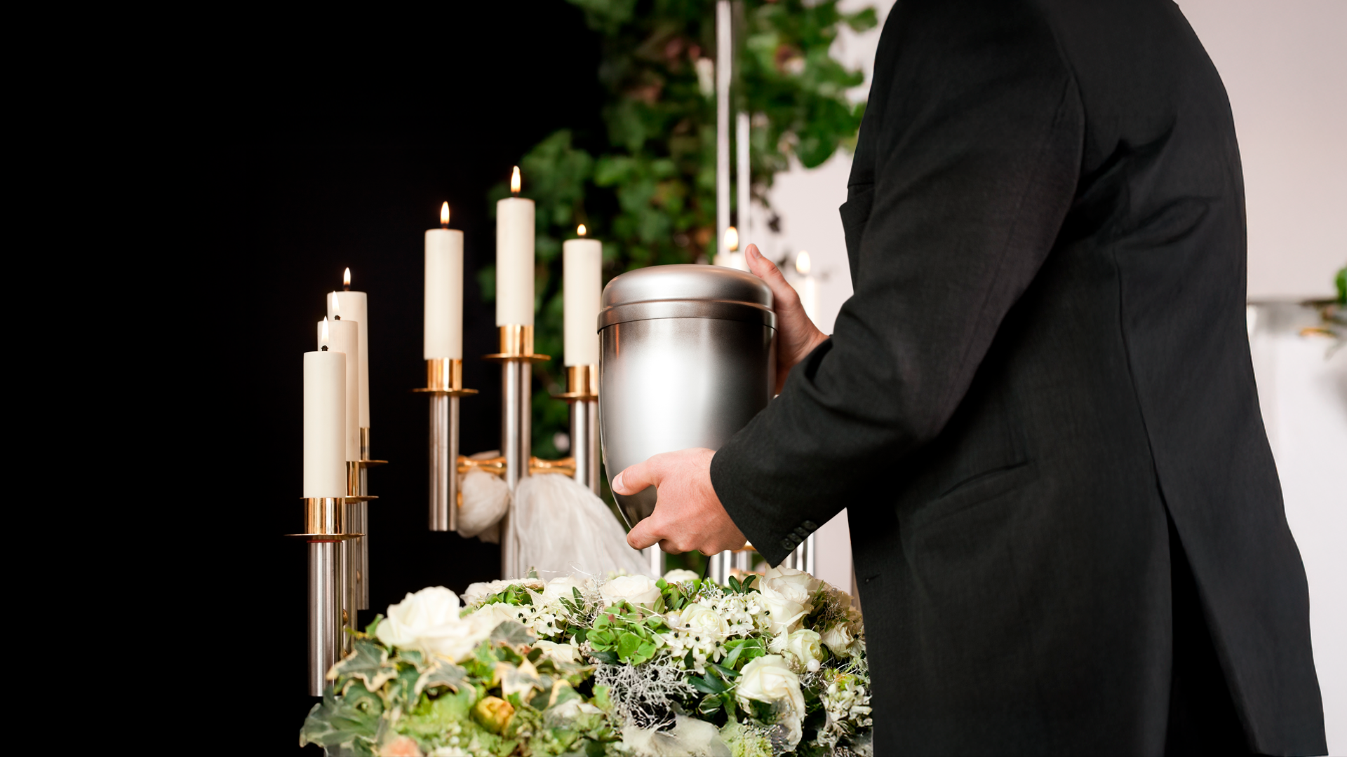 Imagen ilustrativa. Hombre en traje formal sostiene urna funeraria en velorio. Especial del Dia de los fieles difuntos 2025.